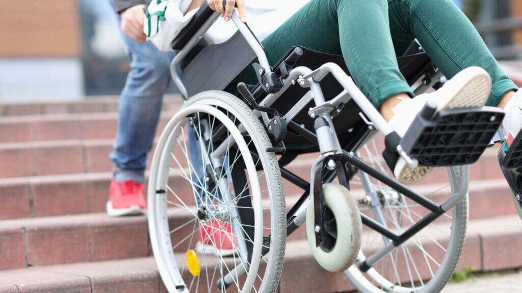 Persona che alza con difficoltà su delle scale una ragazza su una sedia a rotelle