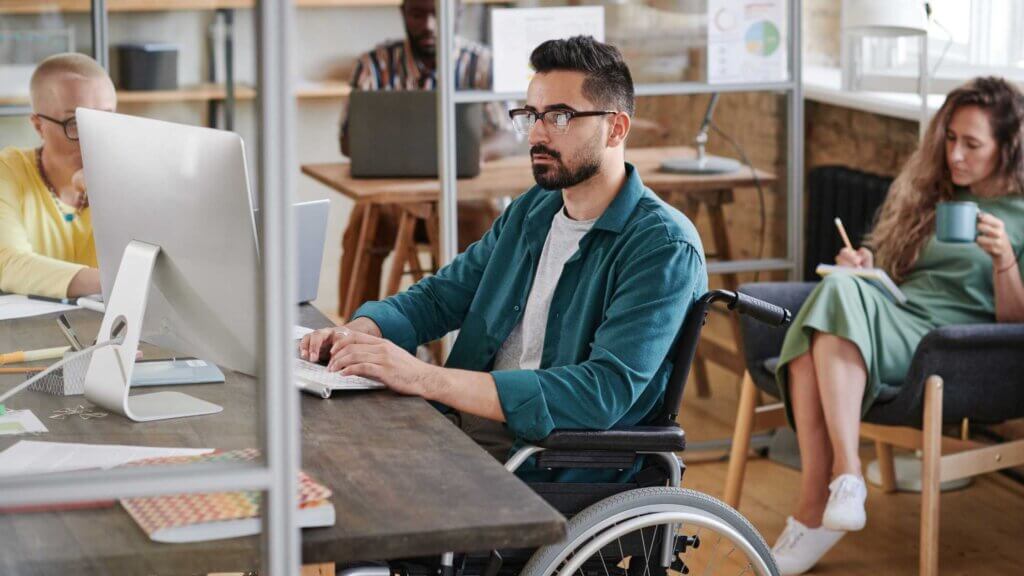 Uomo in sedia a rotelle al computer in ufficio condiviso: inclusione lavorativa e diritti delle persone con disabilità sul posto di lavoro.